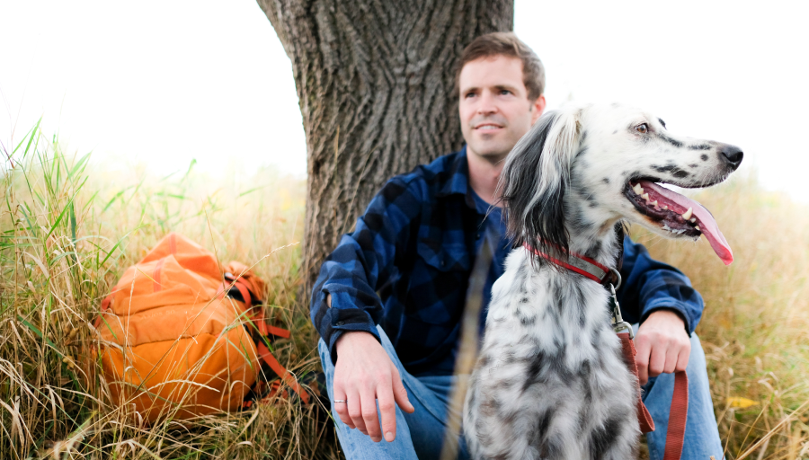 Άντρας κάθεται μπροστά από ένα δέντρο με το πορτοκαλί σακίδιο του στο γρασίδι με τον σκύλο του μπροστά του